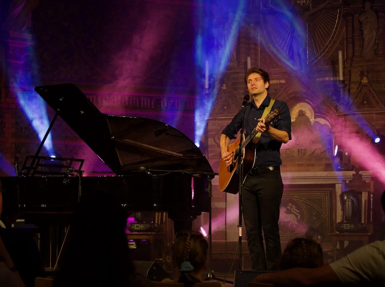 Portrait des Künstlers Lennert Schilgen mit Gitarre in bunt beleuchteter Kirche