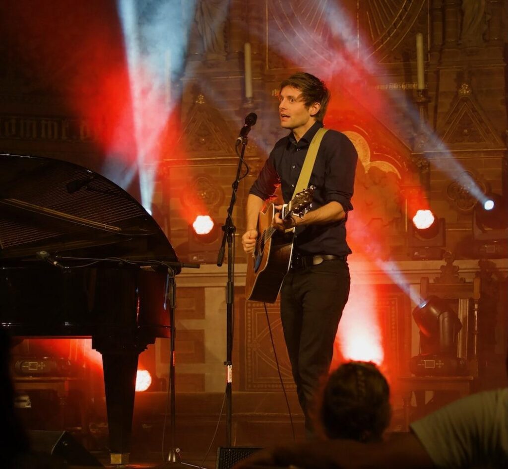 Portrait des Künstlers Lennert Schilgen mit Gitarre in bunt beleuchteter Kirche
